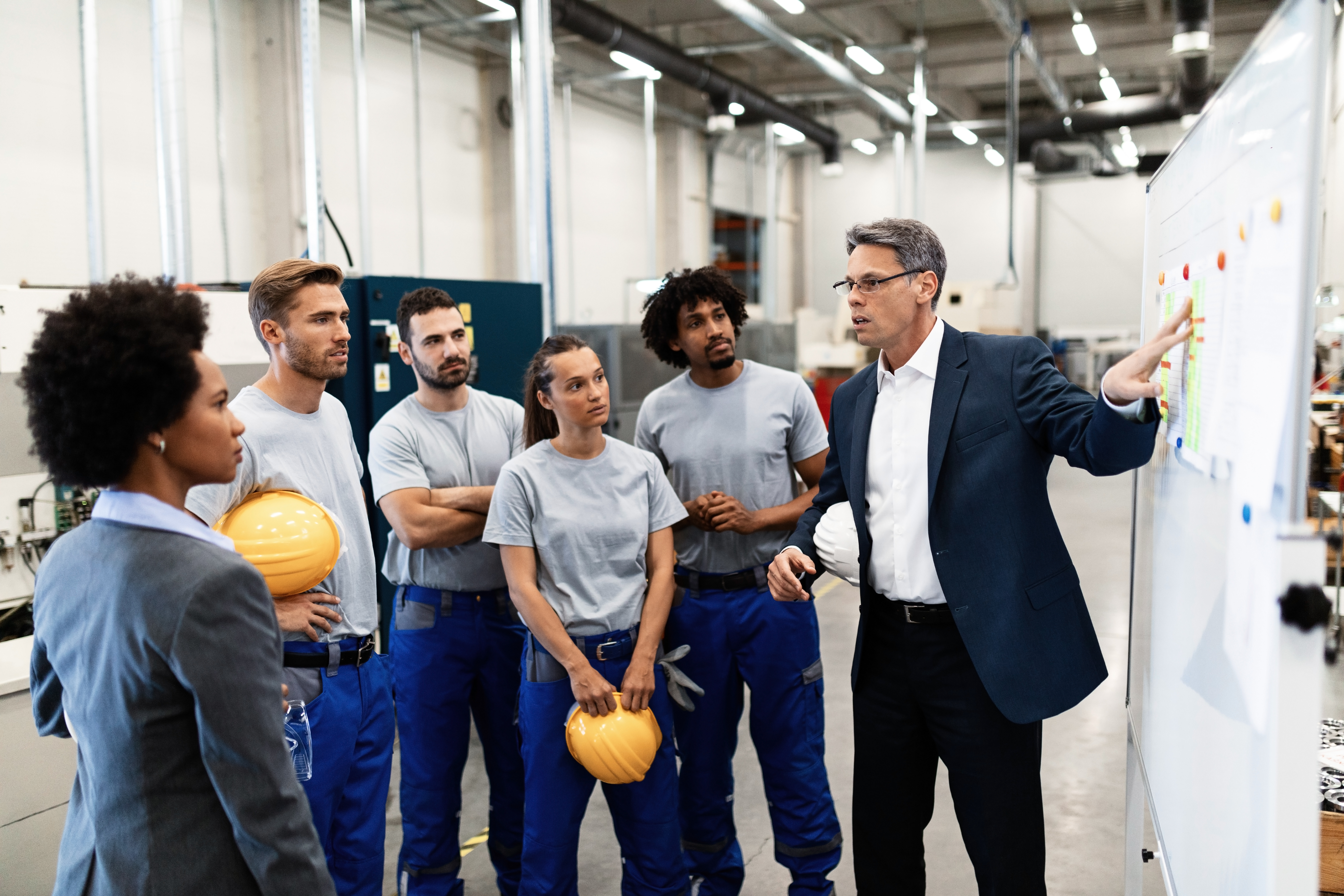 Mature businessman explaining new strategy plans to group of employees in a factory. team watching a presentation